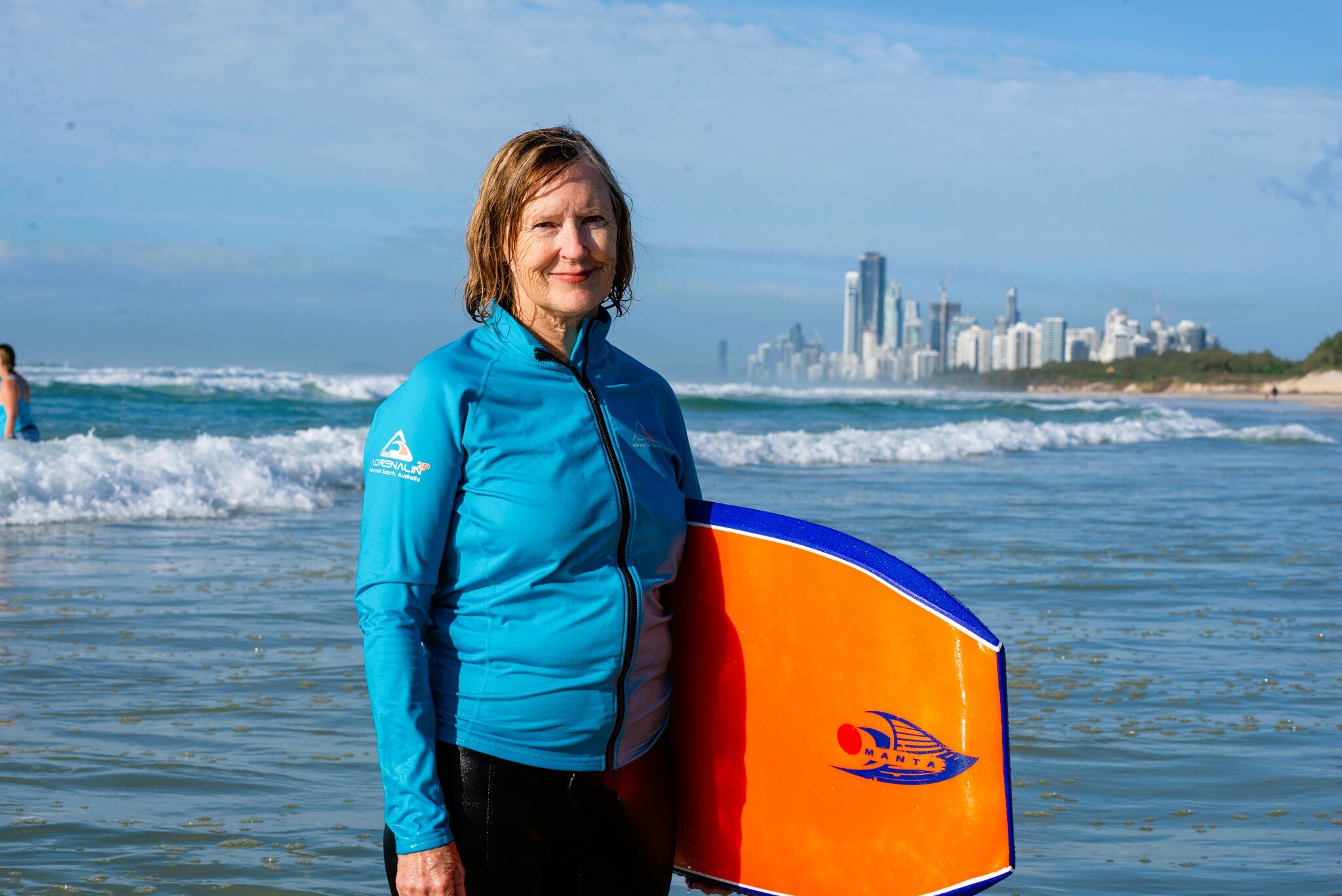 A woman in a long-sleeved, blue, water shirt holding an orange boogie board at the beach.