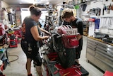 Three people work on motorbike in workshop.