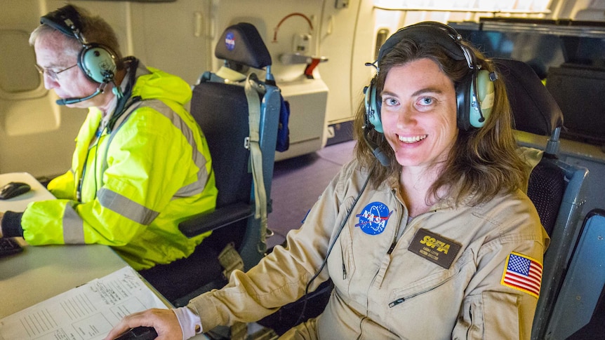 A woman sits at a computer and smiles at the camera, wearing a NASA jumpsuit and a radio headset.