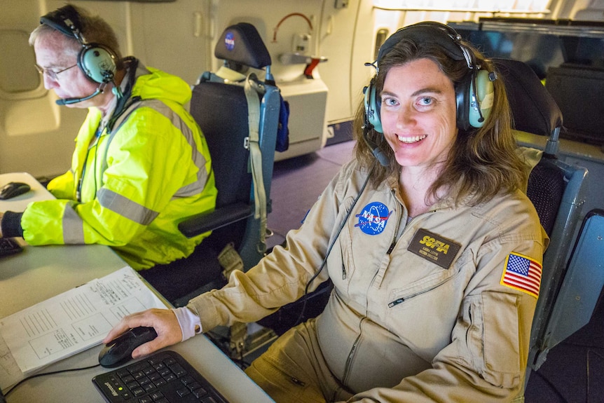 A woman sits at a computer and smiles at the camera, wearing a NASA jumpsuit and a radio headset.