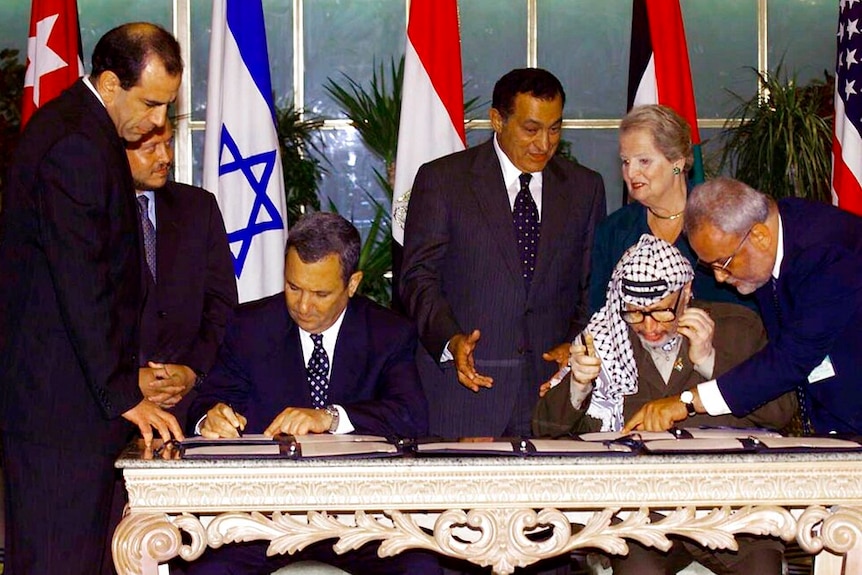 On an ornate table, you view Palestinian and Israeli leaders signing documents as politicians and advisers stand beside them.