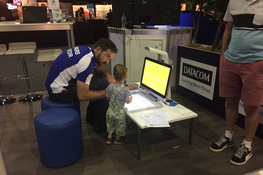 An adult helps a two-year-old boy look at a digital piano and computer.