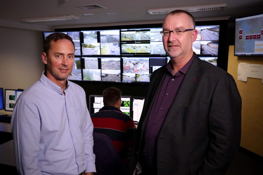 Two men stand in front of traffic monitoring screens, another man sits with his back to the camera