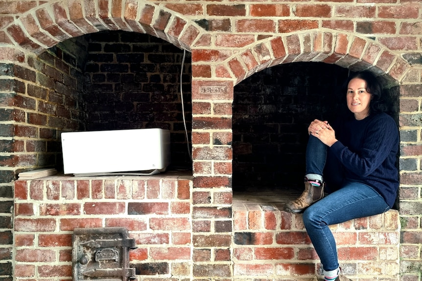 A casually dressed woman sitting comfortably inside a large, old-looking brick fireplace.