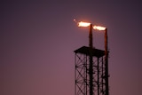 The silhouette of a steel lattice gas tower at dusk with a flare at the top