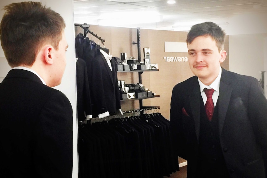 teenage boy stands looking at himself admirably in a mirror while dressed in a suit.