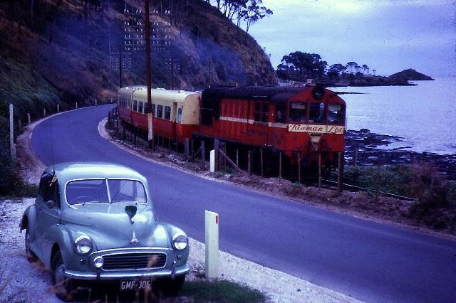 A red train travels beside the beach with a blue car parked in the foreground