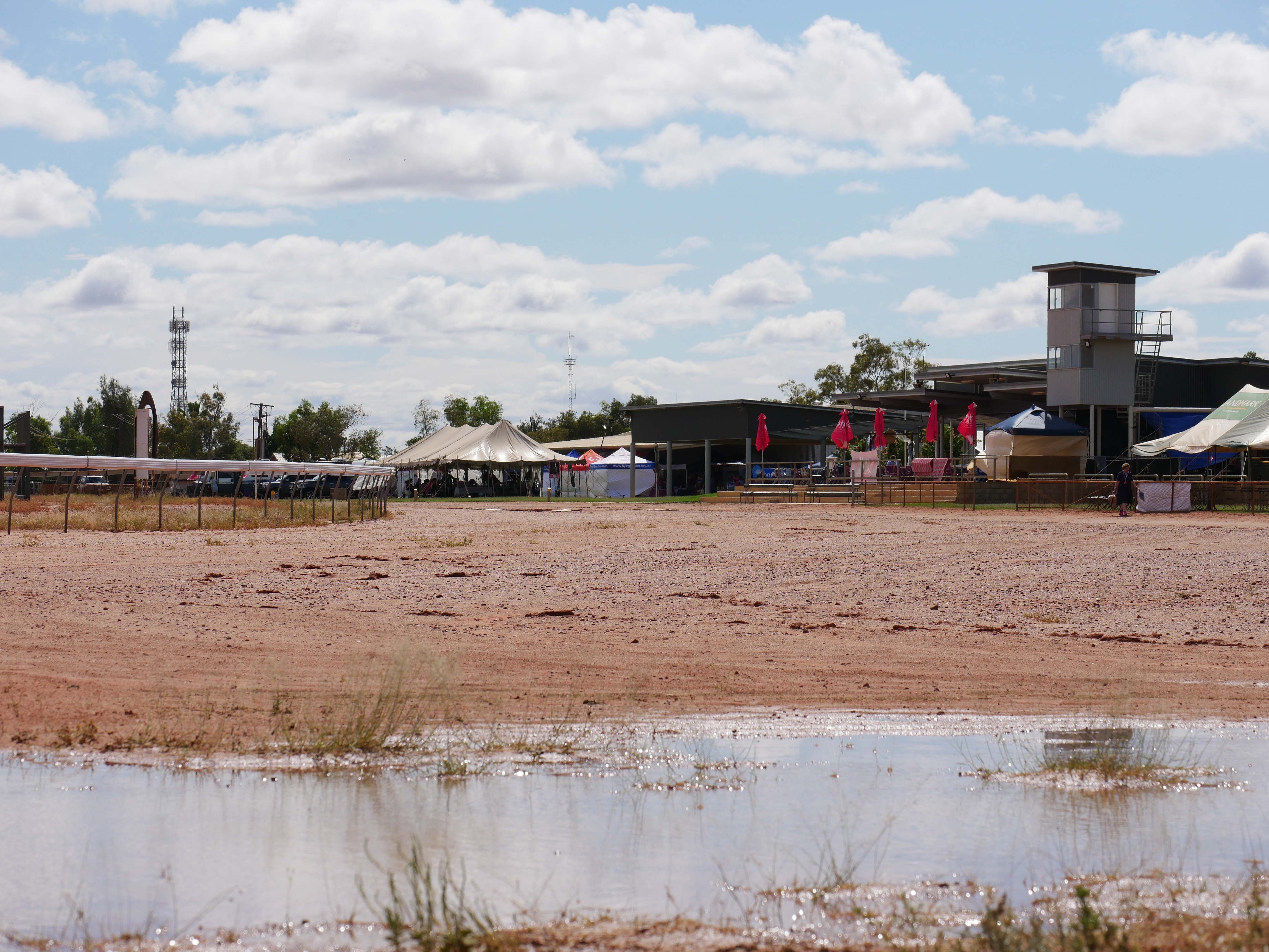 A large puddle at a red dirt racetrack under partly cloudy skies.