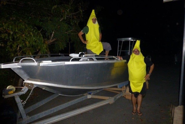 two men dressed in banana suits pose in an aluminium boat on a trailer.