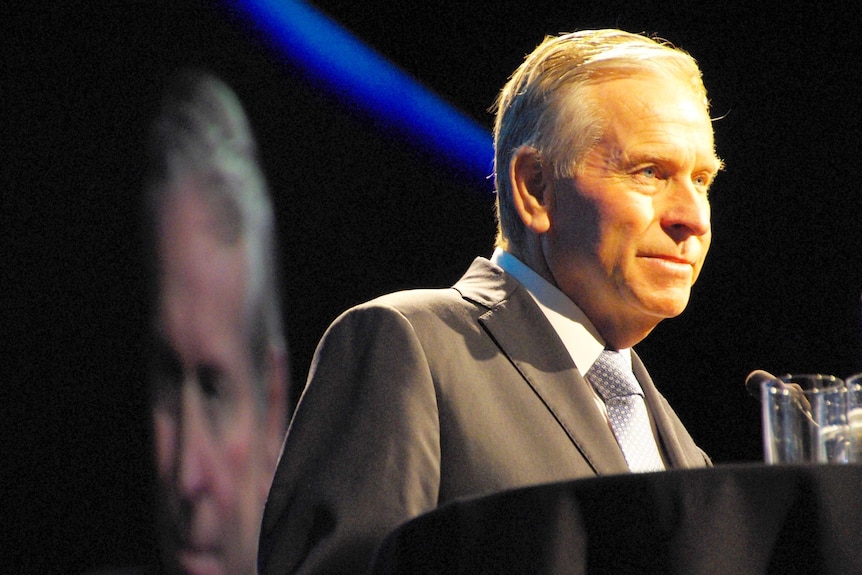 Premier Colin Barnett stands at a podium with a big screen behind him showing his face.