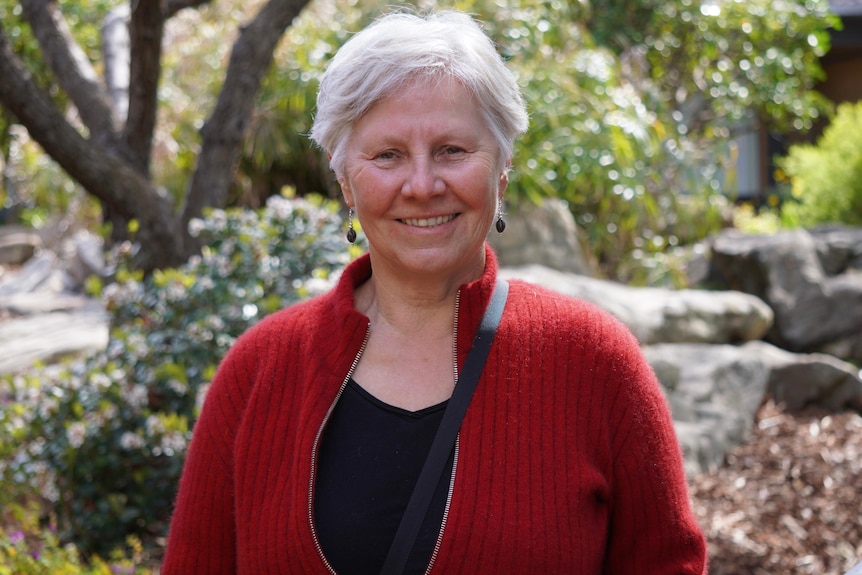 A woman with white hair and a red jumper smiles, standing in front of a garden setting