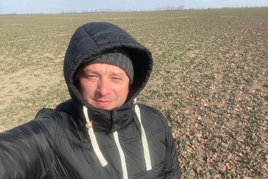 a man in a hoodie and beanie takes a selfie photo in a crop of low lying pasture