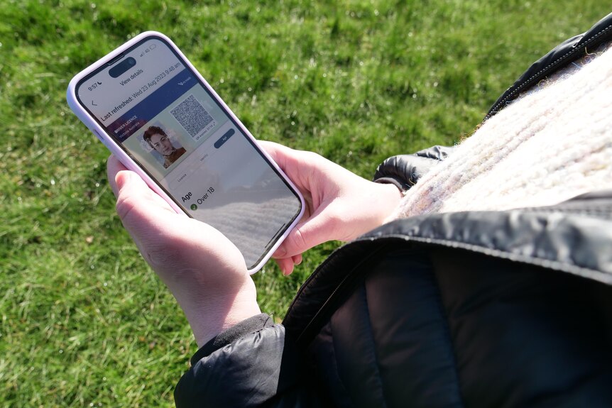 A person, face unseen, stands outside on some grass and operates a phone.