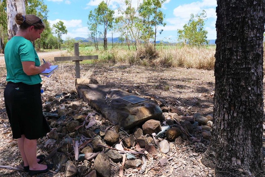 A woman stands beside a lone grave piled under rocks near a farm.