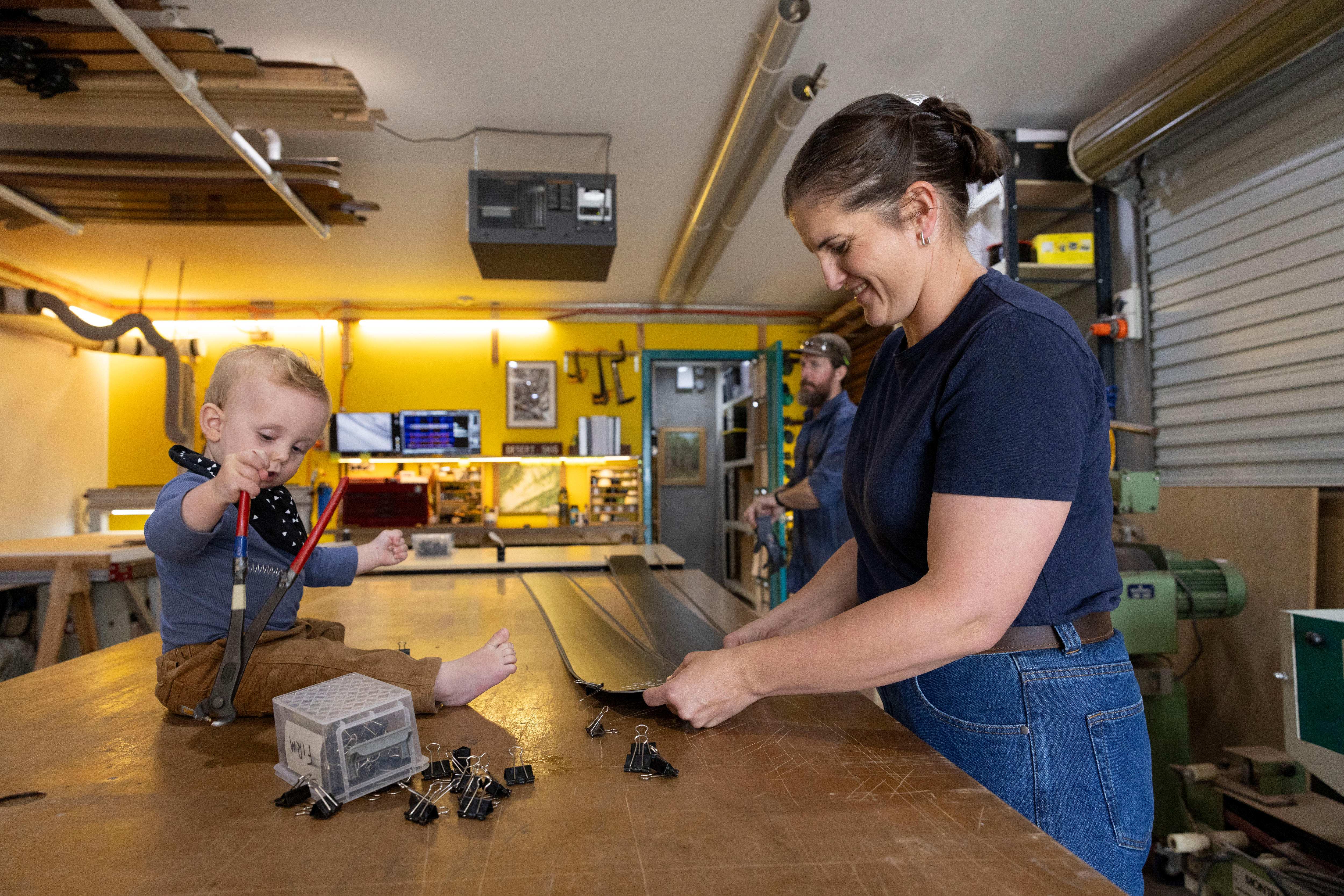 A baby sitting on a bench, with his mother nearby. 