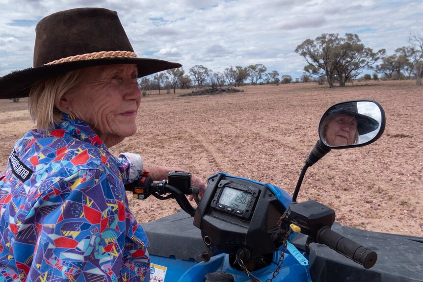 An elderly woman wearing an Akubra hat sits on a quad bike in a dry, dusty paddock.