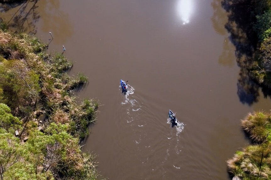 A drone shot from above of two kayakers on a creek with brown-coloured water and vegetation along the banks visible.