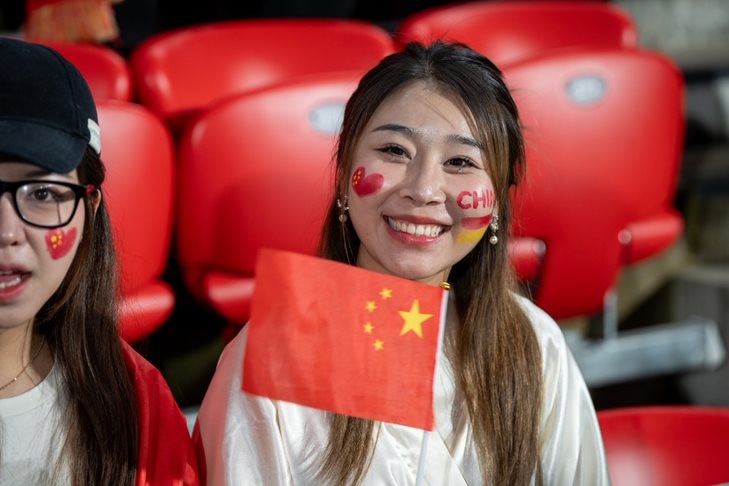 Two young woman sit in the grandstand smiling, with their faces painted, while one holds a Chinese flag in her hands.