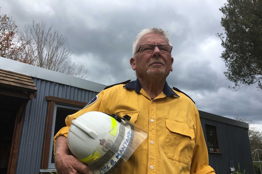 An older man in fire gear stands in front of a house with his helmet under his arm.