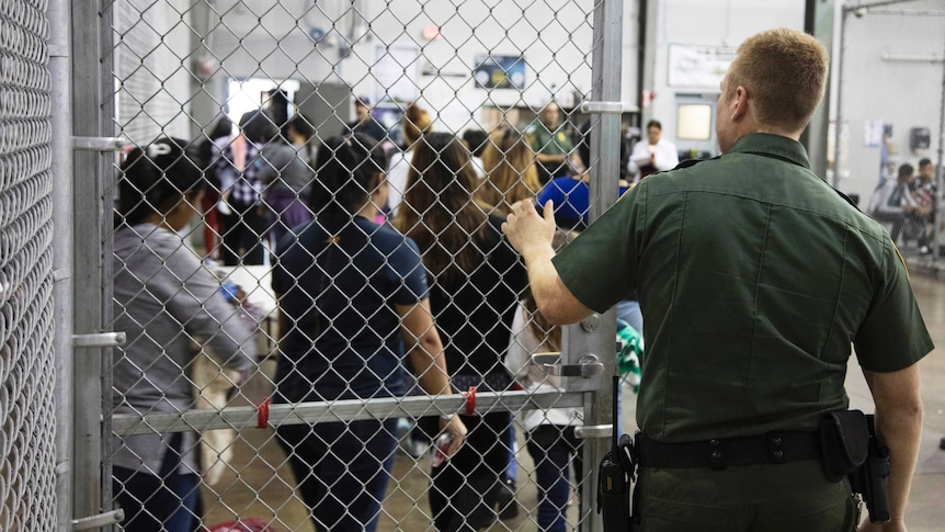 A Border Patrol agent holds a gate open as kids walk out of a cage.