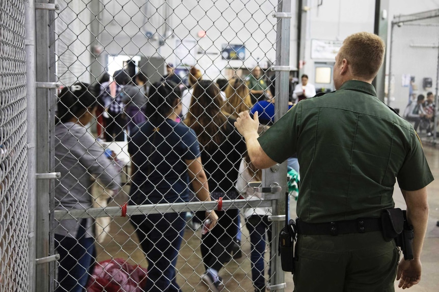 A Border Patrol agent holds a gate open as kids walk out of a cage.