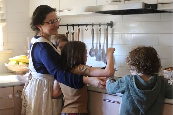 A smiling woman wearing apron stands at kitchen bench with a baby and two older children who are helping to cook.