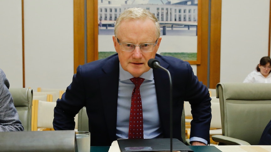 Lowe is wearing a blue suit, looking straight into camera, sitting at a desk.