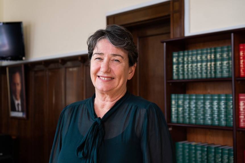 A smiling middle-aged woman with short, dark hair stands in front of a bookshelf.