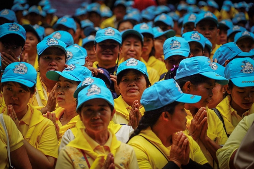 The frame is filled with South East Asian people dressed in yellow shirts and blue caps pressing their palms together.