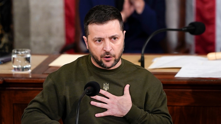 VOlodymyr Zelenskyy, wearing a khaki jumper, sits at a desk while holding one hand to his chest