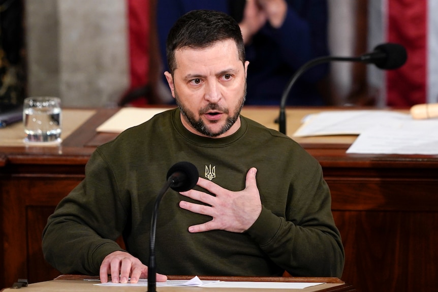 VOlodymyr Zelenskyy, wearing a khaki jumper, sits at a desk while holding one hand to his chest