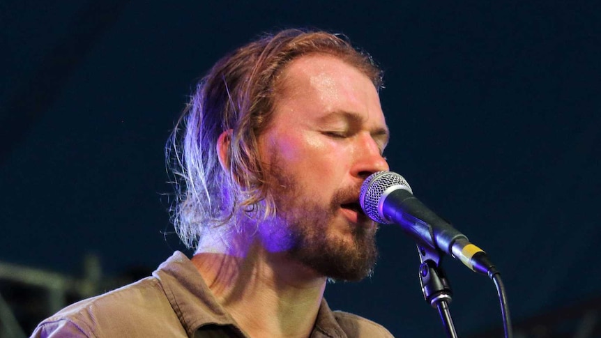 John Teskey sings into a microphone with his eyes closed, holding a guitar