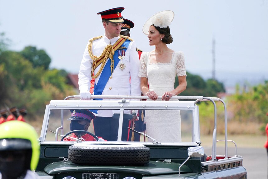 Prince William in a Royal Naval dress uniform and Kate Middleton in a white dress stand in the back of an open jeep