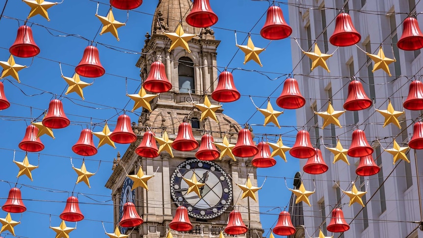 You look up at a grid of street Christmas decorations on a clear blue day, with a classical clock tower behind it.