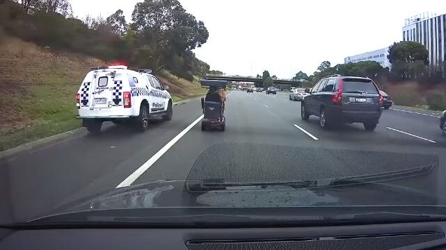 A police car with lights flashing drives next to a mobility scooter on a freeway.