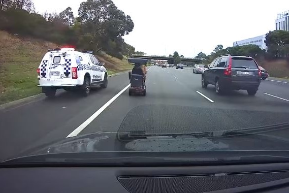 A police car with lights flashing drives next to a mobility scooter on a freeway.