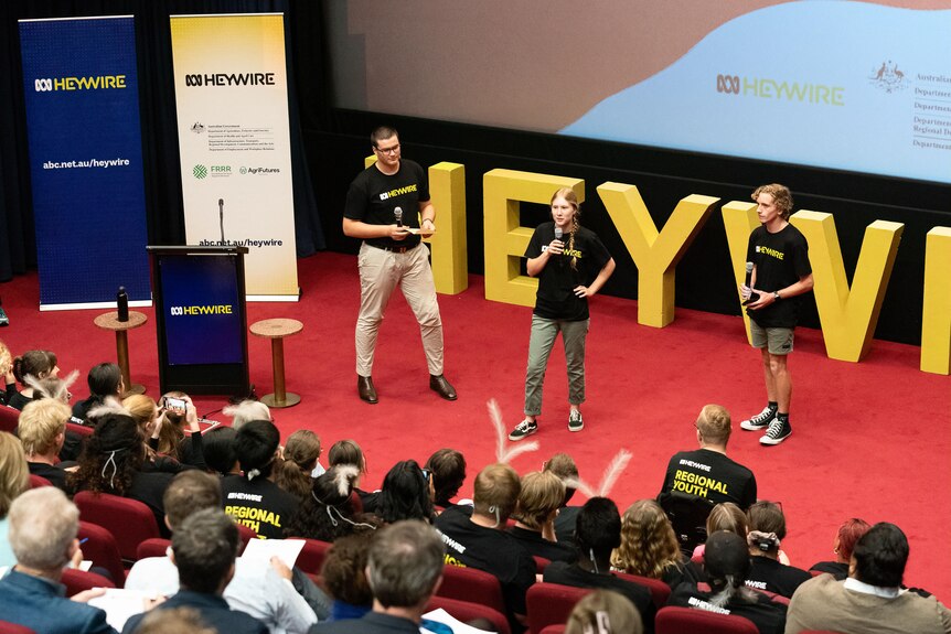 Three people stand on a stage with two banners saying Heywire on their left side. Middle holds mic up to mouth