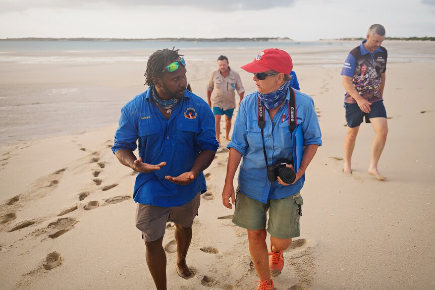 A male ranger and female scientist, followed by several other people, speaking while walking along an otherwise empty beach.