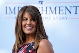 Monica Lewinsky smiles and looks over her shoulder while standing in front of a promotion for Impeachment.