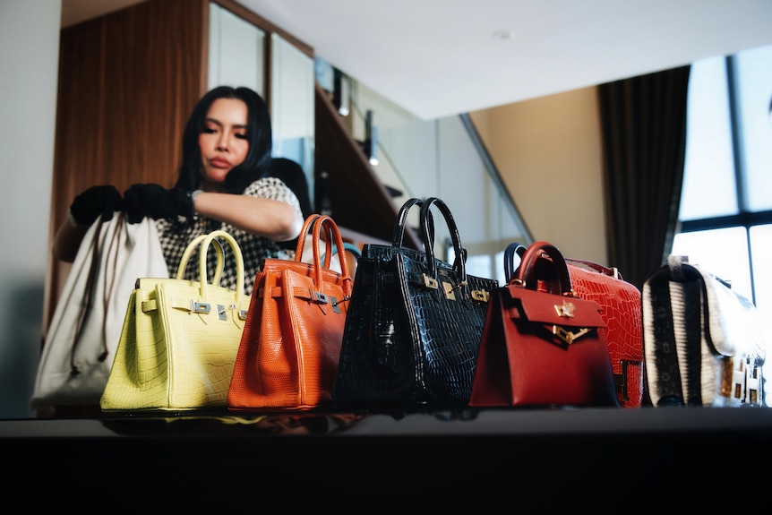 A woman wearing black gloves places a handbag down alongside five others in a row: yellow, orange, black, red, beige