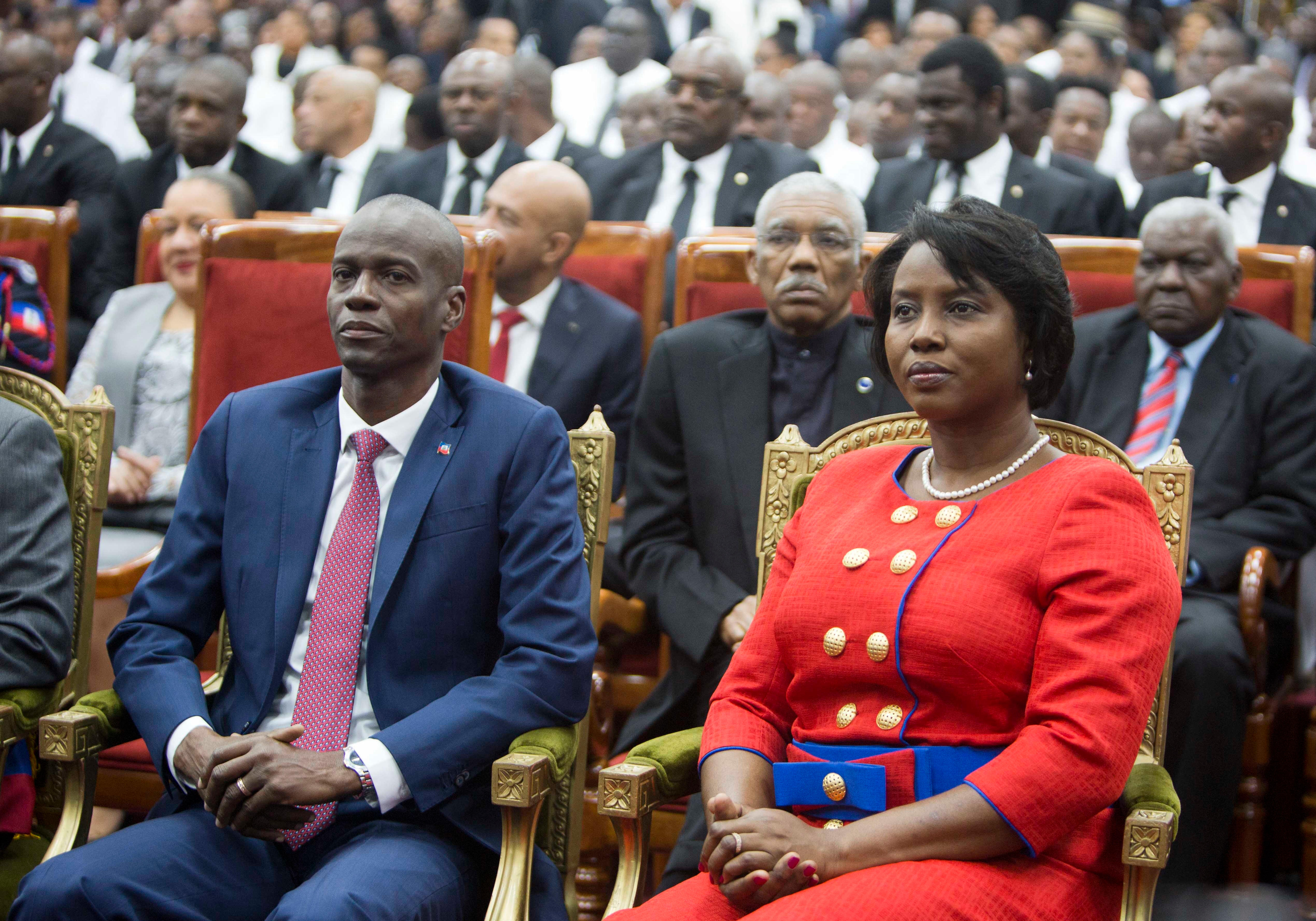 A middle-aged Haitian couple in tailored clothes sit in gold-trimmed chairs at the front of a seated crowd.