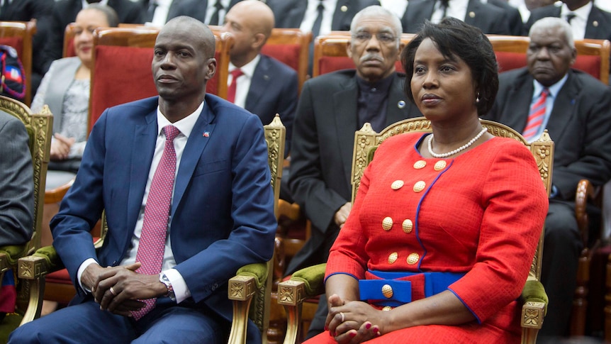A middle-aged Haitian couple in tailored clothes sit in gold-trimmed chairs at the front of a seated crowd.