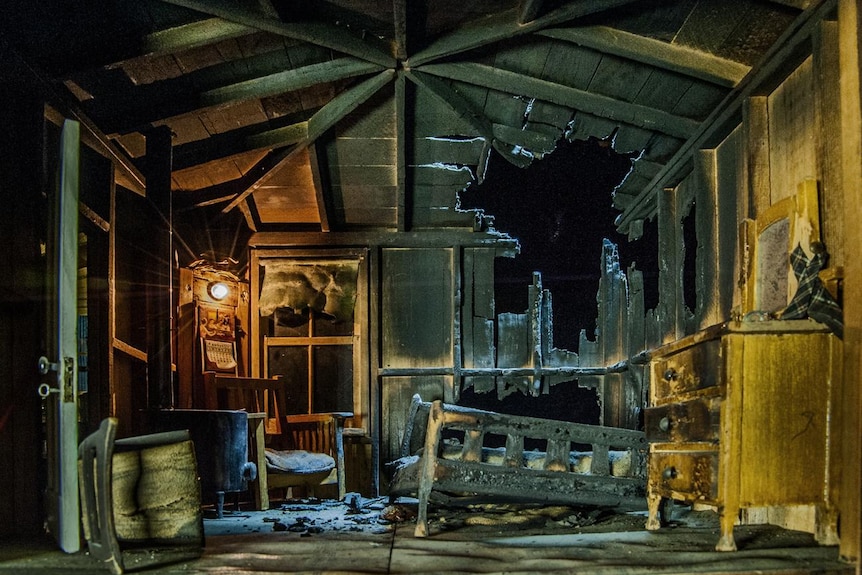 The interior of a house, with burn wooden roof panels, charred wooden furniture and overturned chairs.