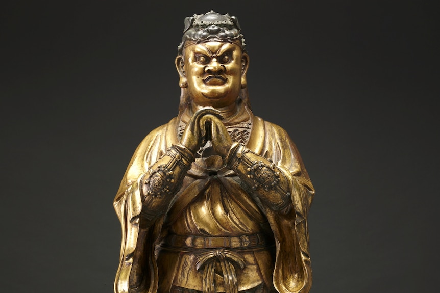 A golden, bronze coloured statue of a Chinese guardian holding hands clasped in fists under his chin.