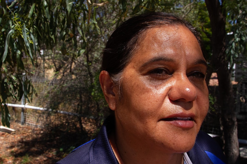A close-up of an Aboriginal woman's face bathed in dappled sunlight as she stands under a tree in a backyard.