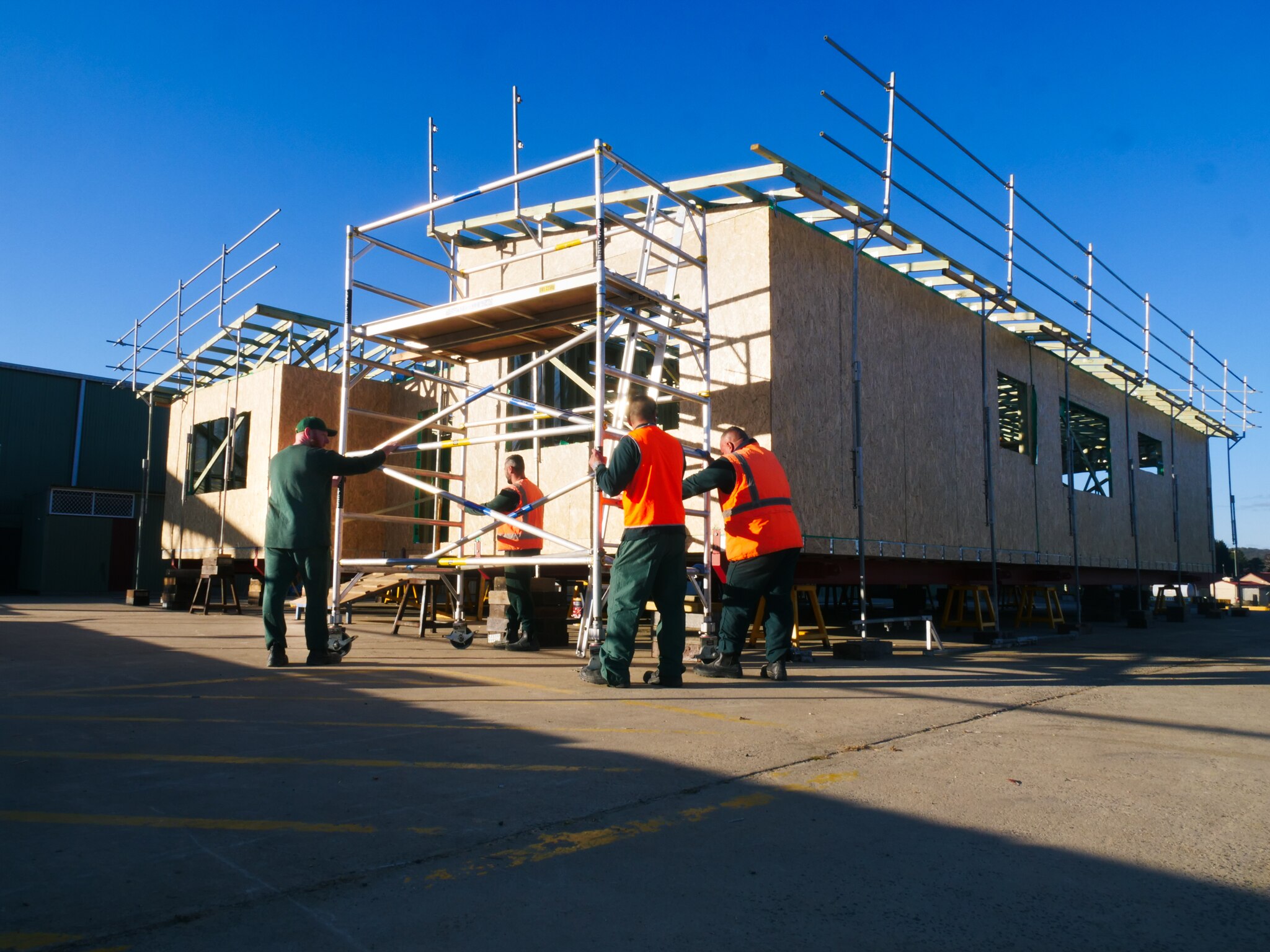 Men in prisoner greens move scaffolding in front of home under construction.