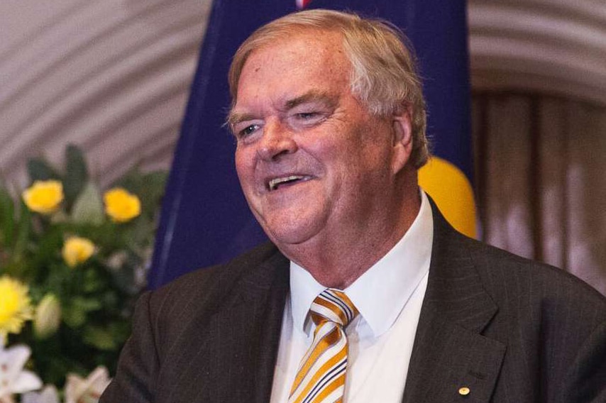 A head and shoulders shot of a smiling Kim Beazley standing at a podium in front of a flag and flowers.