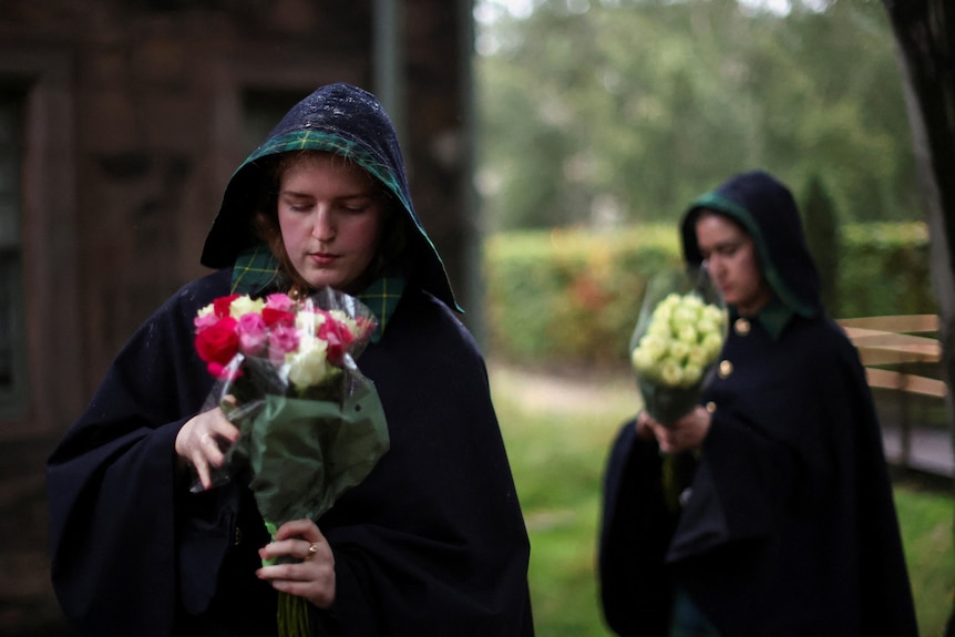Two women dressed in black and green cloaks carry flowers. 