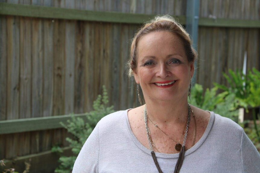 A middle-aged woman with silver earings and neckless smiles at camera in suburban backyard.