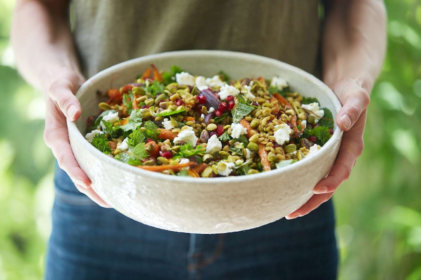 Two hands hold a serving bowl of grain salad for a celebration, with freekeh, pistachio, goat cheese, roasted carrots and herbs.